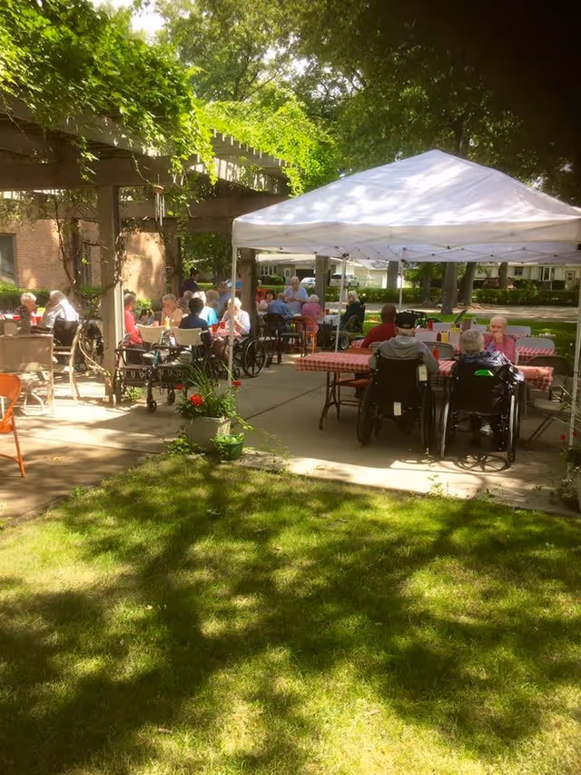 Outdoor gathering at Robbinswood Northcrest Campus with elderly residents seated at tables under a white canopy and in the open air, surrounded by greenery and shaded by trees.