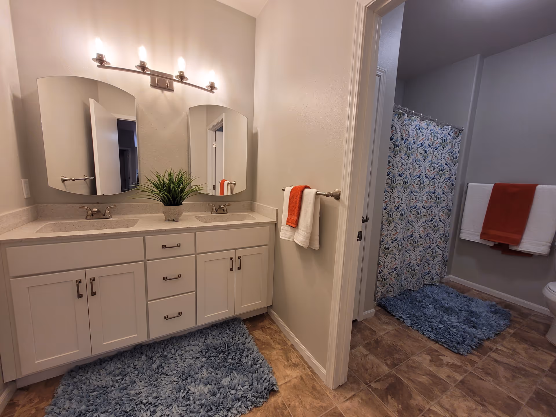 A bathroom with a double sink vanity featuring white cabinets and two mirrors above. There is a small green plant placed between the sinks. The floor has brown tiles with two blue shaggy rugs. A doorway leads to a shower area with a colorful patterned shower curtain and a towel rack holding white and red towels.