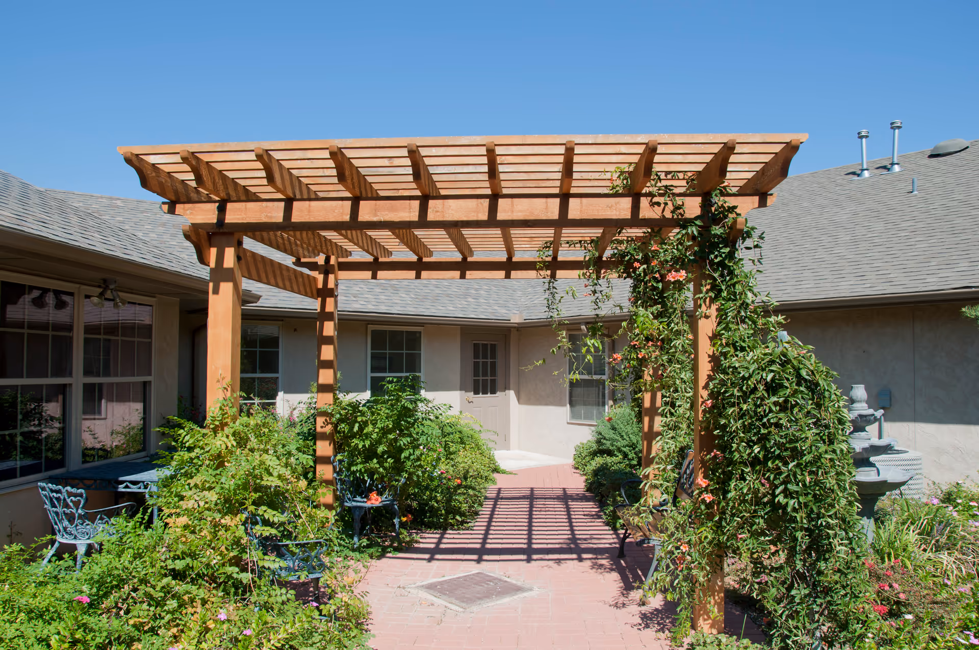 Outdoor courtyard area at Autumn Hill Manor featuring a wooden pergola with climbing plants, surrounded by green bushes and flowers, with a brick pathway and a water fountain in the background under a clear blue sky.