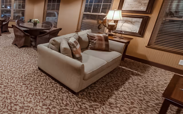 A cozy living room area with a beige sofa adorned with two plaid pillows, a side table with a lamp, and framed artwork on the wall. In the background, there are round tables with wicker chairs and a window with blinds.