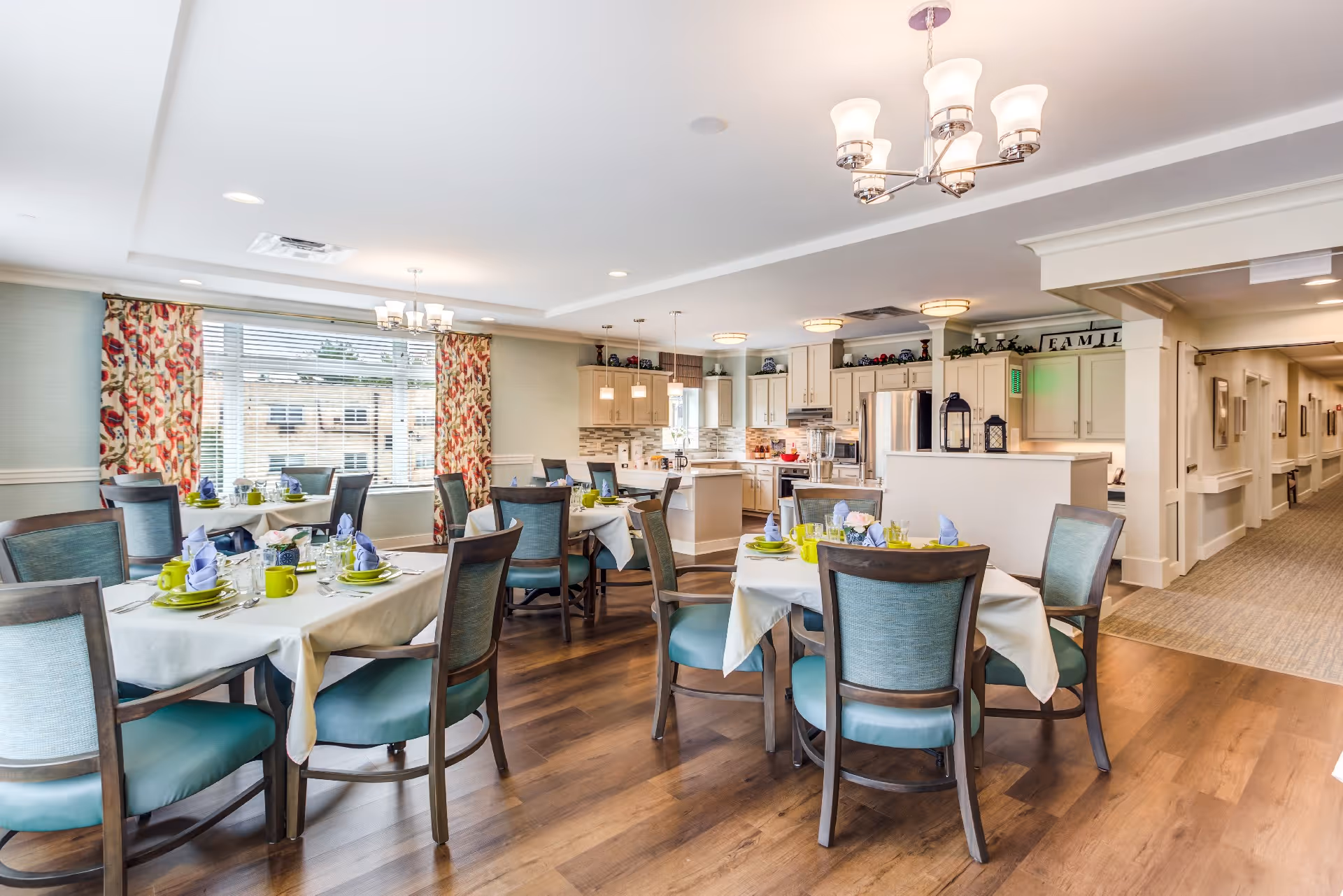 Bright communal dining room with round tables set for meal service and an open kitchen in the background.