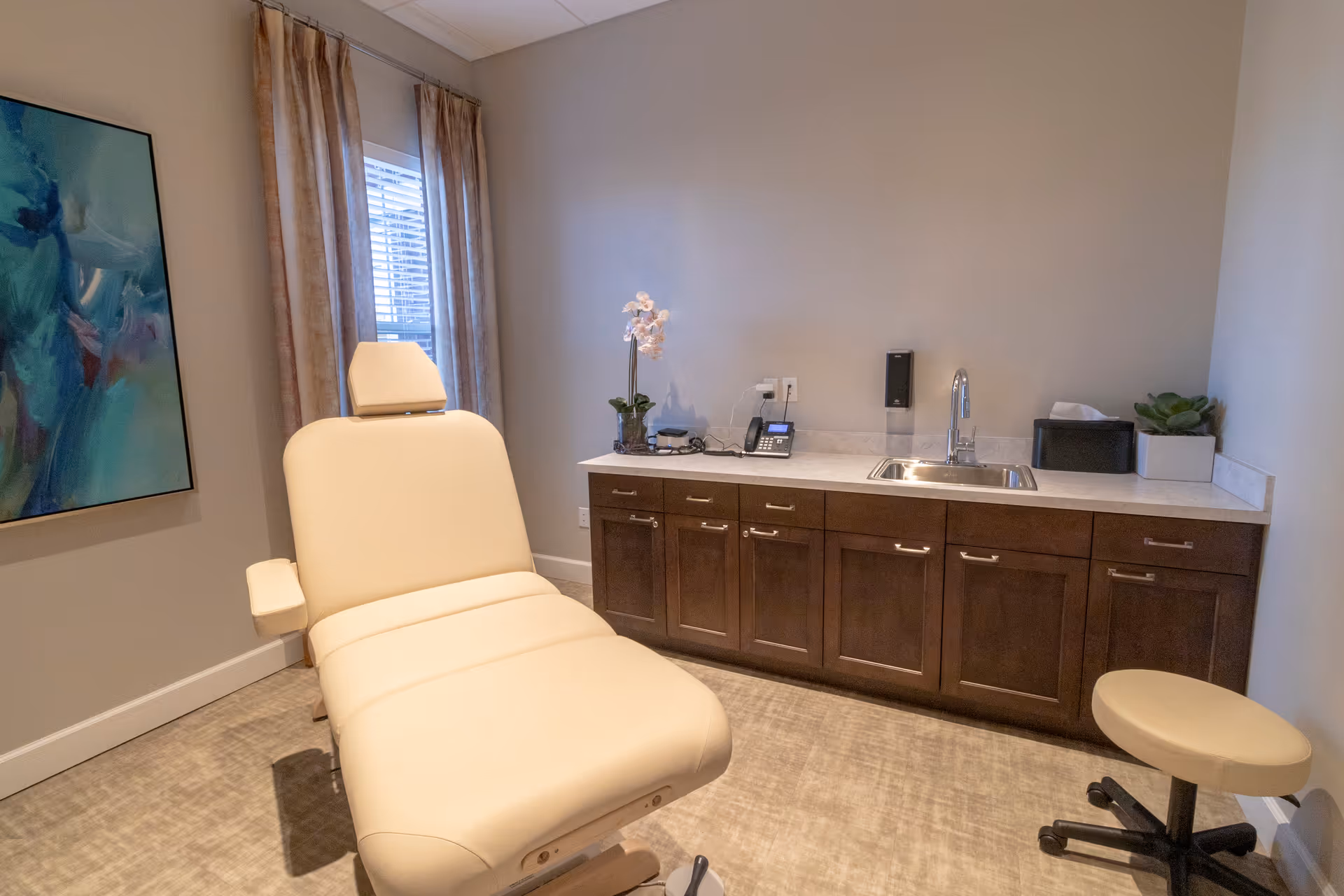 A small medical or therapy room with a beige reclining chair in the center, a rolling stool to the right, and a countertop with a sink, telephone, tissue box, and potted plants along the back wall. There is a window with curtains and a large abstract painting on the left wall.