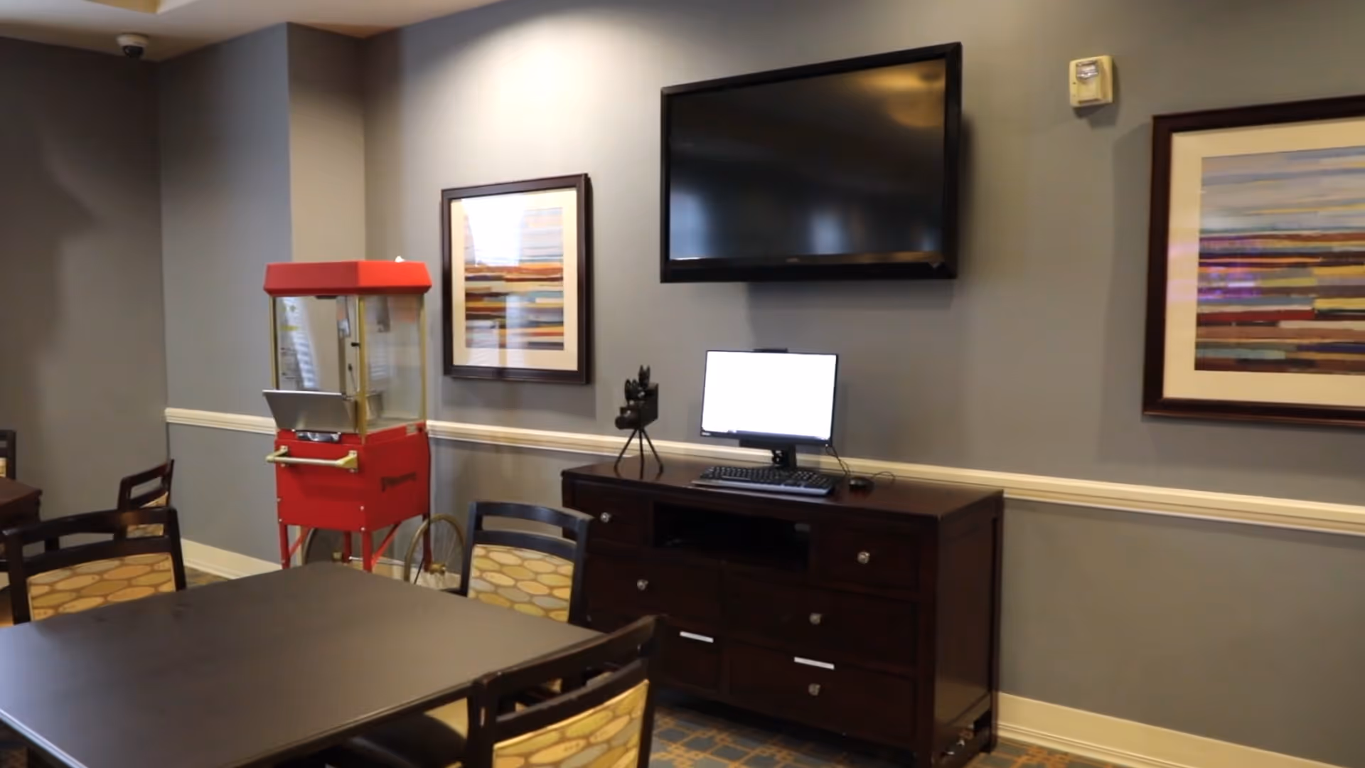 Interior room with a dark wooden table and chairs with patterned cushions, a red popcorn machine on wheels, a dark wooden cabinet with a computer monitor and keyboard on top, a wall-mounted flat screen TV, and two framed abstract paintings on a gray wall.