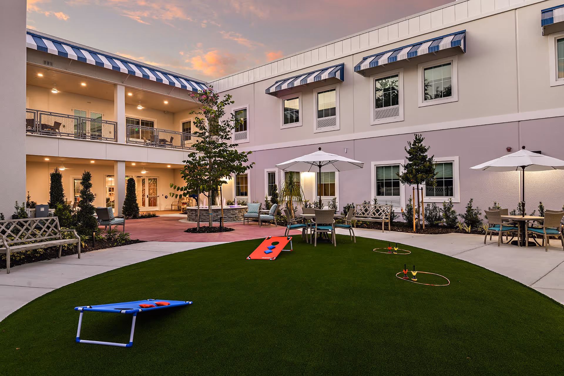 Outdoor courtyard of a senior living facility with artificial turf, lawn games, seating areas and umbrellas in front of a two-story building with balconies.