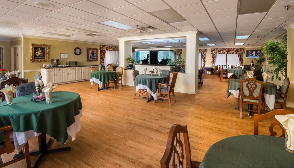 A spacious dining room with several round tables covered with green and white tablecloths, each set with folded napkins and small floral centerpieces. The room features wooden chairs, a large fish tank built into a partition wall, floral curtains on the windows, framed artwork on the walls, and a television mounted in the corner. The floor is wooden, and there is a counter area with a coffee maker and microwave along one wall.