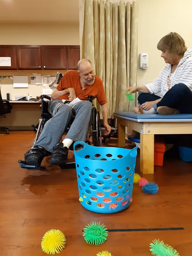 An elderly man in a wheelchair and a woman sitting on a padded table engage in a playful activity with colorful spiky balls and a blue plastic basket in a room with wooden flooring and beige curtains.