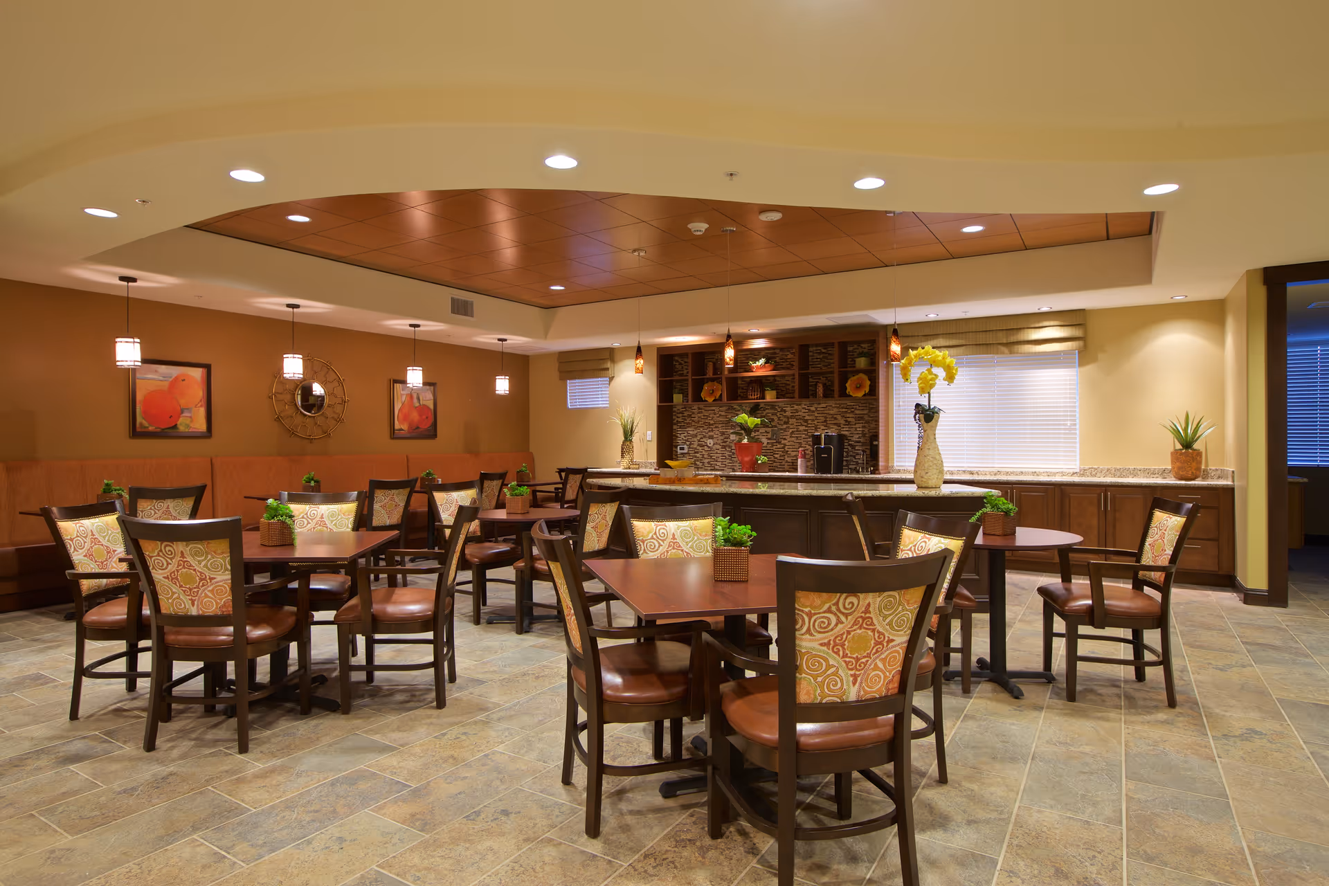 A well-lit dining area with several wooden tables and chairs featuring patterned upholstery. The room has a tiled floor, a ceiling with recessed lighting, and pendant lights hanging above the tables. There are small potted plants on each table and decorative artwork on the walls. In the background, there is a counter with cabinets, a coffee machine, and more plants, creating a warm and inviting atmosphere.