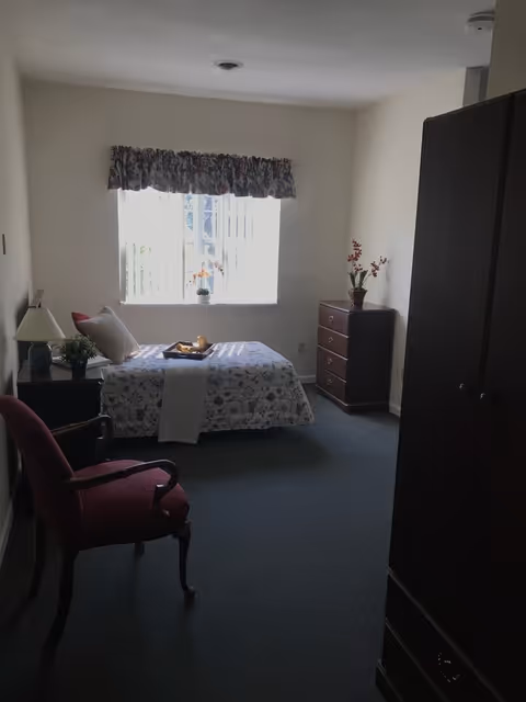 A simple bedroom with a single bed covered with a floral bedspread and a white runner. There is a wooden nightstand with a lamp and a small plant next to the bed. A wooden chest of drawers with a potted plant is against the far wall. A window with vertical blinds and a floral valance lets in natural light. A red upholstered armchair is positioned near the foot of the bed, and a dark wooden wardrobe is visible on the right side of the room.