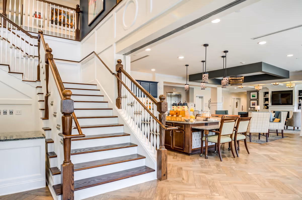 Bright and spacious interior of a senior living facility featuring a wooden staircase with white railings, a central island with chairs, pendant lights hanging from the ceiling, and a comfortable seating area with armchairs and a television in the background.