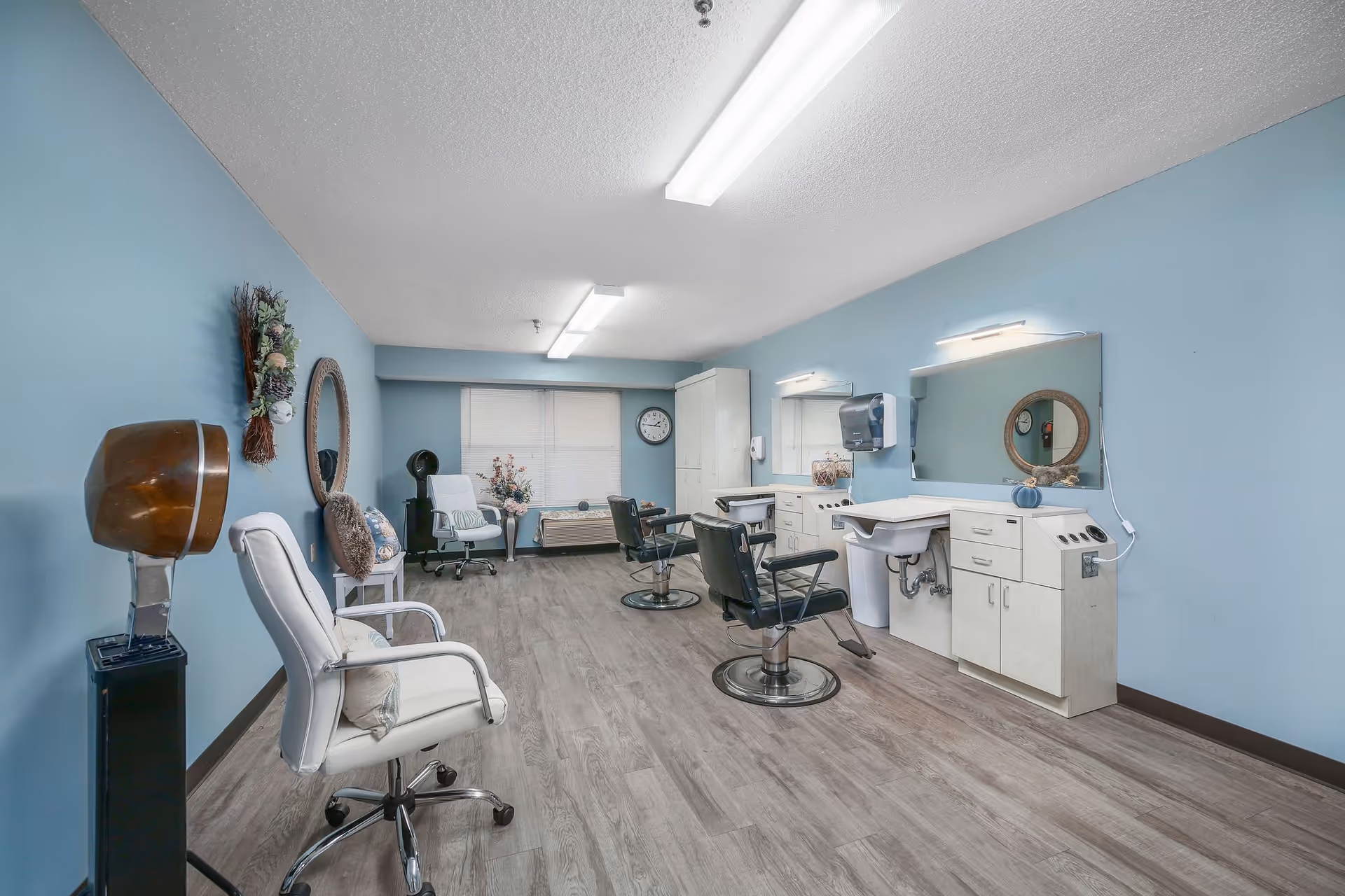 Interior of a salon room with light blue walls and wood flooring. The room contains two black salon chairs in front of sinks and mirrors, a white swivel chair, a vintage hair dryer, a round wall mirror, and decorative items including a wreath and flowers. Fluorescent ceiling lights illuminate the space.