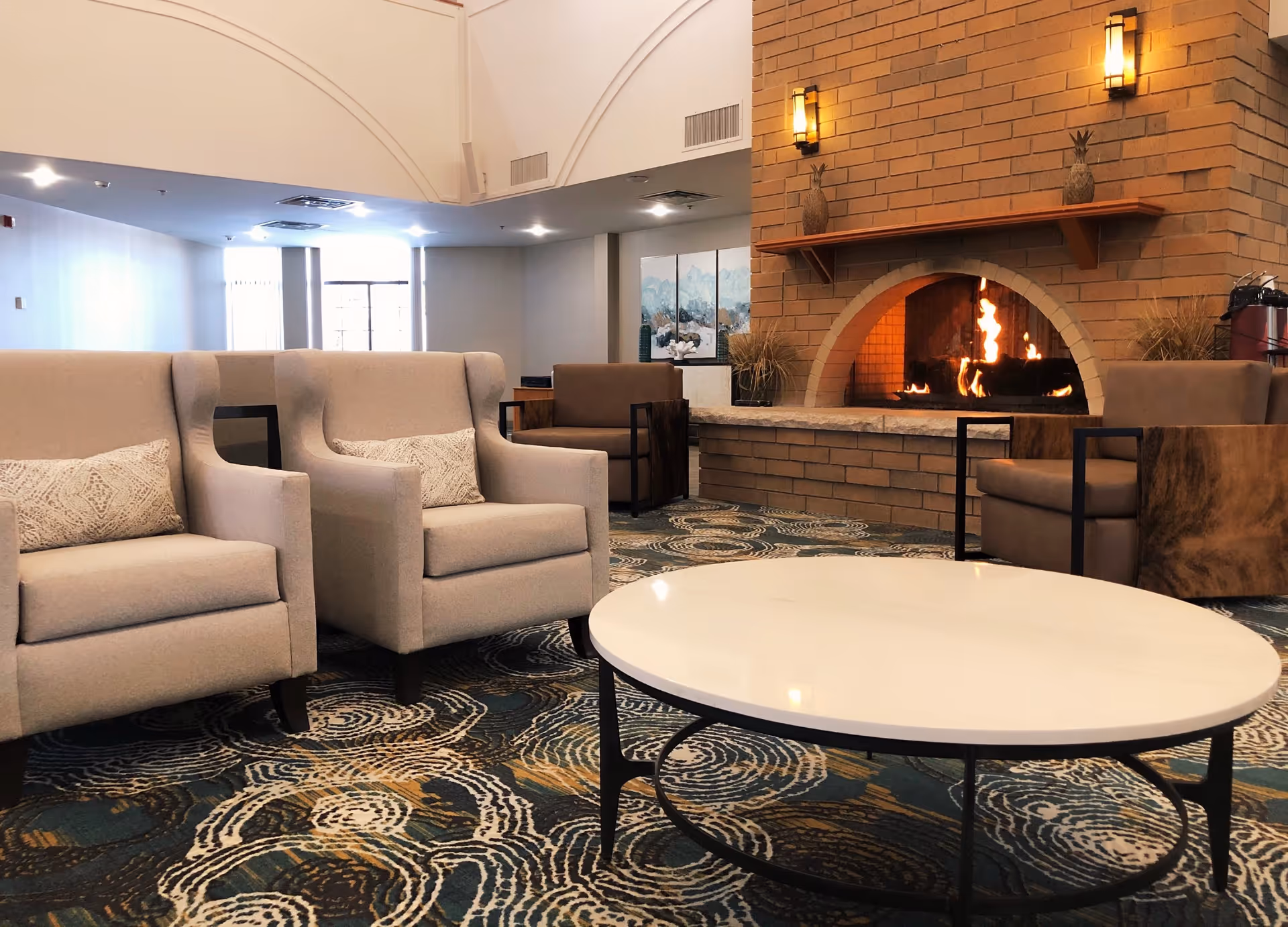 A cozy living room area with a lit brick fireplace, two beige armchairs with patterned cushions, two brown armchairs, a round white coffee table, and a patterned carpet. The room has soft lighting and a calm, inviting atmosphere.