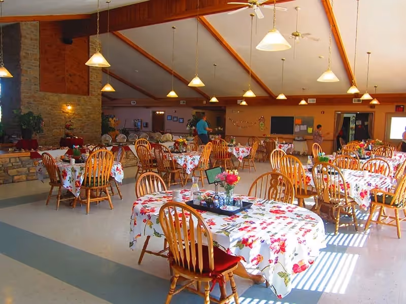 A bright dining room with multiple wooden tables and chairs. The tables are covered with floral tablecloths and decorated with small flower arrangements and condiments. The room has a high ceiling with wooden beams and hanging pendant lights. There are a few people in the background near the walls and a stone fireplace on the left side.