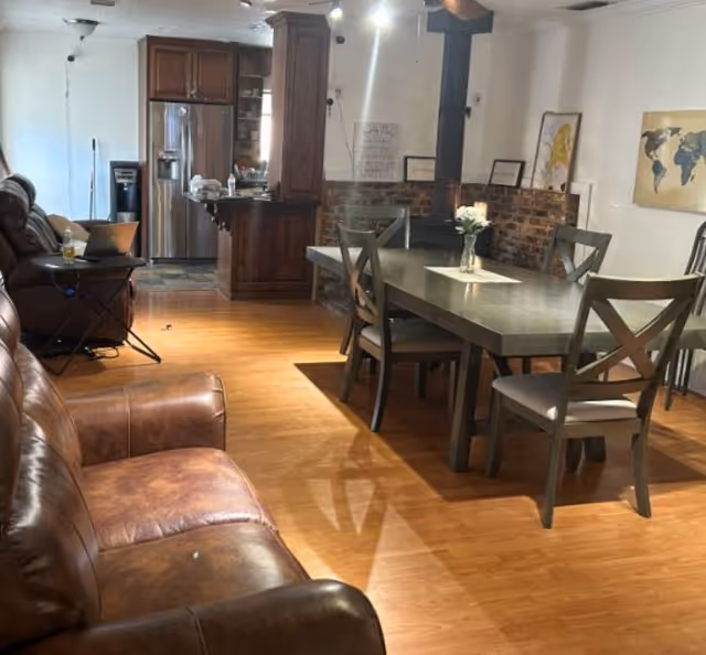 Open-plan living area with leather couches, a dining table and chairs, and a kitchen with stainless refrigerator in the background.