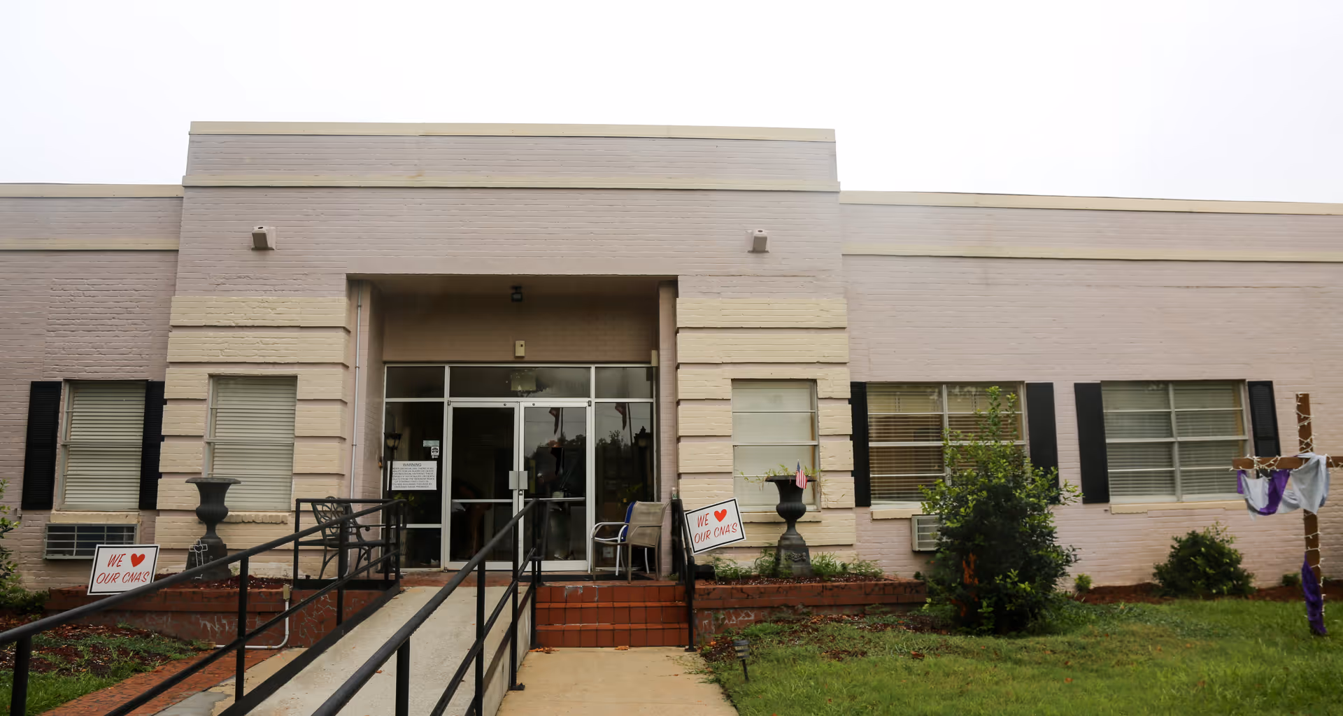 Front exterior view of a single-story building with a ramp and stairs leading to glass double doors. The building has light-colored brick walls with black window shutters and several windows. There are two signs near the entrance that say 'WE ♥ OUR CNAs'. A wooden cross with purple and white cloth is on the right side of the image in the grass.