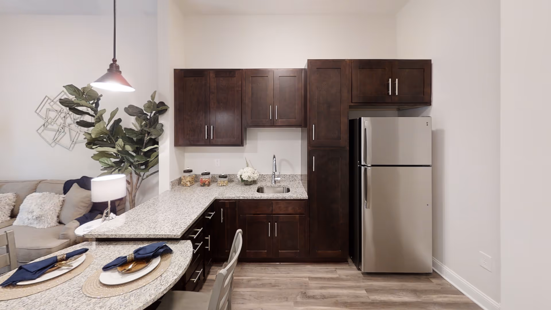Modern kitchen area with dark wood cabinets, a stainless steel refrigerator, granite countertops, a small sink, and a breakfast bar set with plates, gold utensils, and navy blue napkins. In the background, a living room area with a beige couch, pillows, a floor lamp, and a large green plant is partially visible.