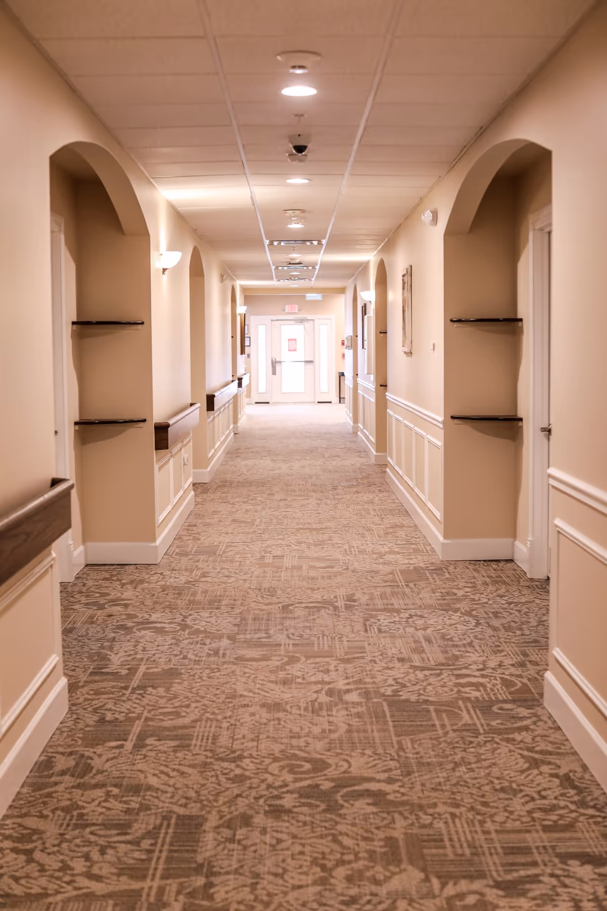 A long, well-lit hallway in a senior living facility with beige walls, patterned carpet, wall-mounted handrails, and arched alcoves with shelves. The hallway leads to a door with windows at the far end.