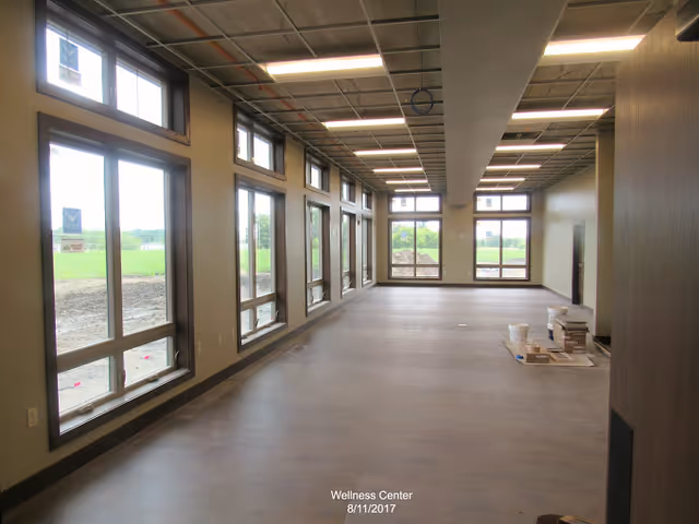 Interior view of a spacious room with large windows along two walls allowing natural light to enter. The room has a high ceiling with exposed grid framework and fluorescent lighting. The floor is covered with wood-like material, and there are some construction materials and supplies stacked on the floor near one wall. Outside the windows, green fields are visible.
