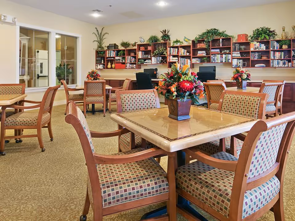 Well-lit common room with square dining tables, patterned chairs, floral centerpieces, and bookshelves along the back wall.