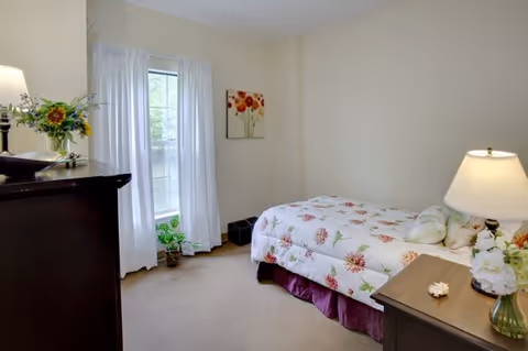A cozy bedroom with a single bed covered in a floral quilt. There is a wooden nightstand with a lamp and a vase of flowers next to the bed. A window with white curtains lets in natural light, and a chest of drawers with a lamp and a flower arrangement is on the left side of the room. A floral painting hangs on the wall.