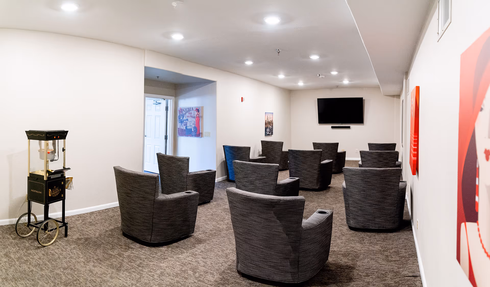 Small screening/common room with gray swivel chairs arranged facing a wall-mounted TV and a popcorn machine near the entrance.