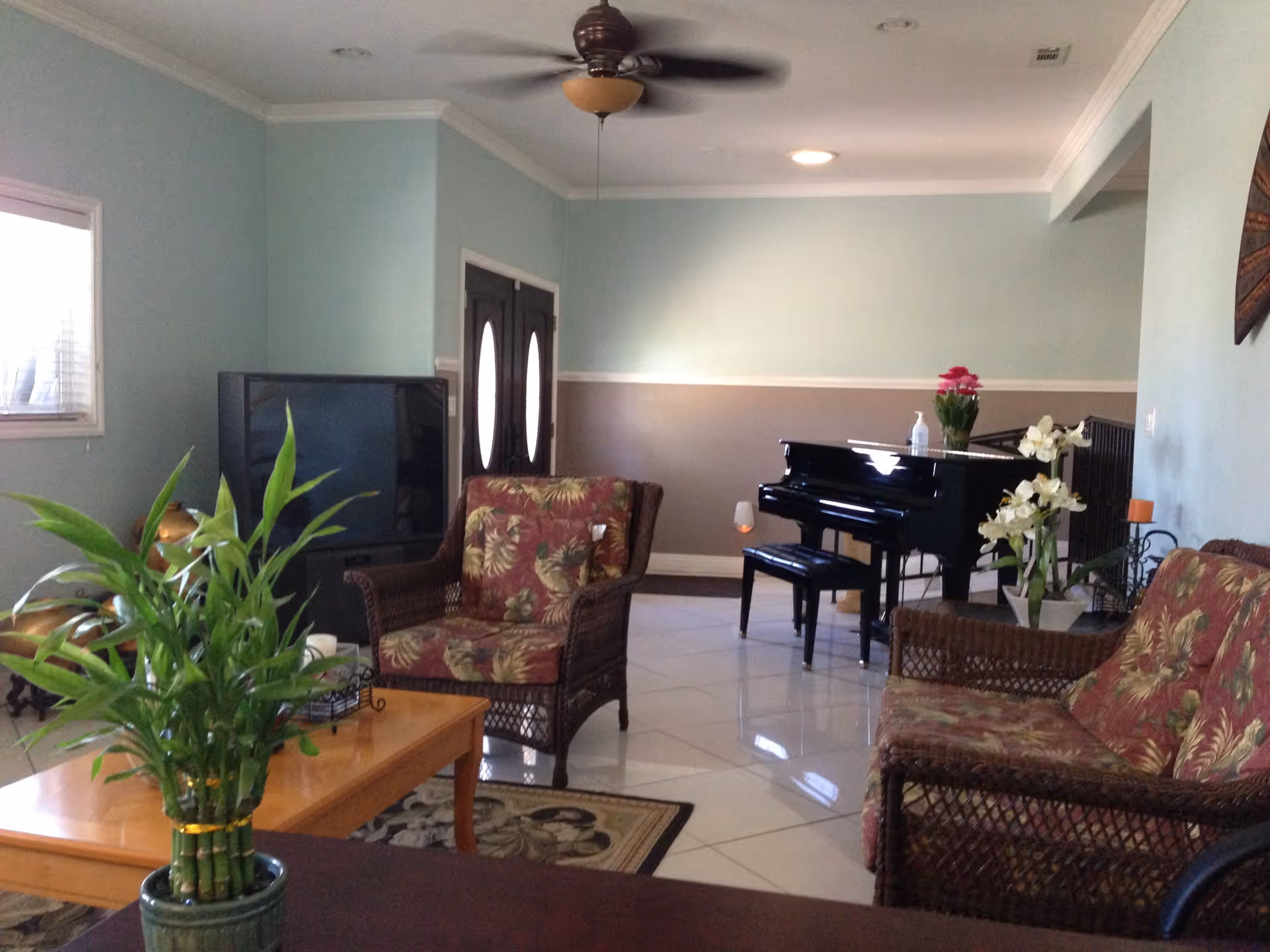 A cozy living room with wicker furniture featuring floral cushions, a wooden coffee table with a potted plant, a large flat-screen TV, a ceiling fan, and a black grand piano with flowers on top. The walls are painted light blue and beige with white trim.