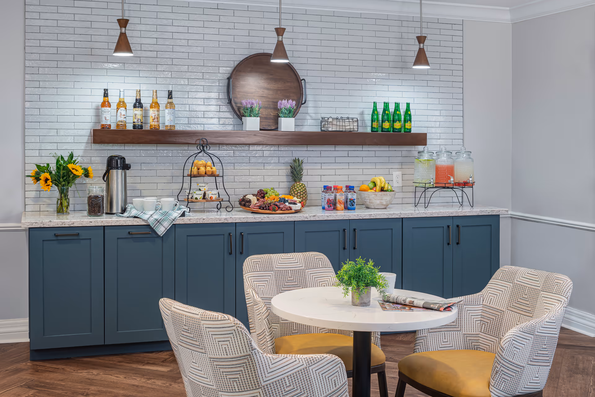 A cozy refreshment area with a white round table surrounded by four patterned chairs with yellow cushions. Behind the table is a countertop with blue cabinets, decorated with a vase of sunflowers, a coffee dispenser, cups, a tiered tray with muffins, a fruit platter, bottled drinks, and three large beverage dispensers with different colored drinks. Above the counter is a wooden shelf holding bottles, two small potted plants, and a large round wooden tray mounted on a white tiled wall. Three pendant lights hang from the ceiling.