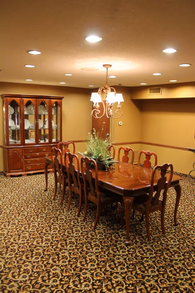 A formal dining room with a long polished wooden table surrounded by eight matching chairs. A decorative floral centerpiece is on the table. The room has patterned carpet flooring, beige walls with a wooden chair rail, and a wooden china cabinet filled with plates and glassware. A chandelier with four lights hangs above the table, and recessed ceiling lights illuminate the room.