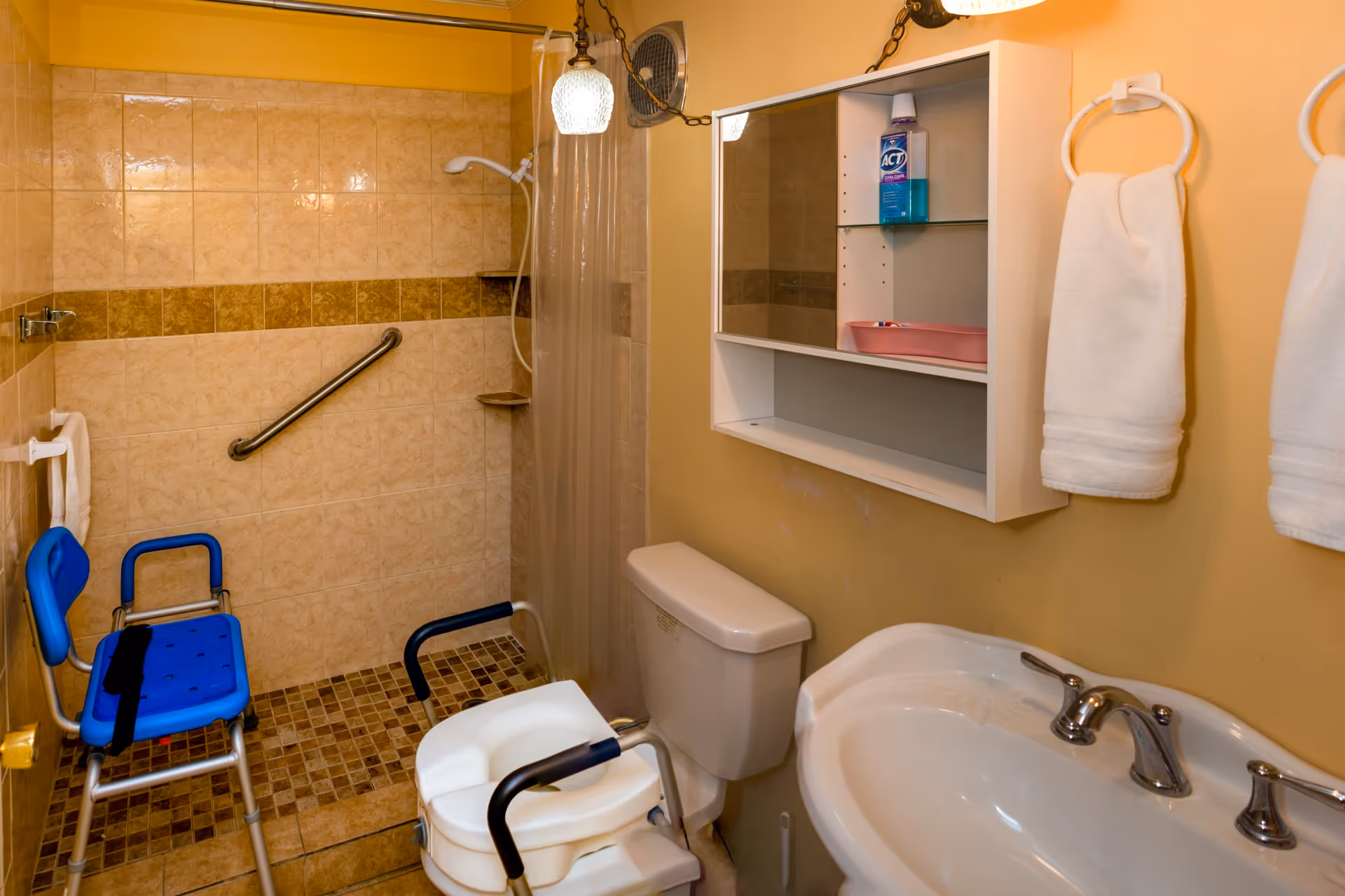 Bathroom with beige tiled shower area equipped with a grab bar and a blue shower chair. There is a white toilet with armrests, a white sink with a silver faucet, a wall-mounted cabinet with a mirror, and two white towels hanging on towel rings. The walls are painted yellow.