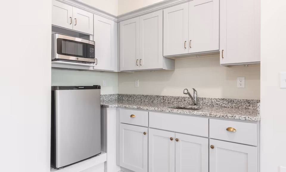 Small kitchen area with white cabinets, a granite countertop, a stainless steel microwave mounted above a compact stainless steel refrigerator, and a single-handle faucet over a small sink.