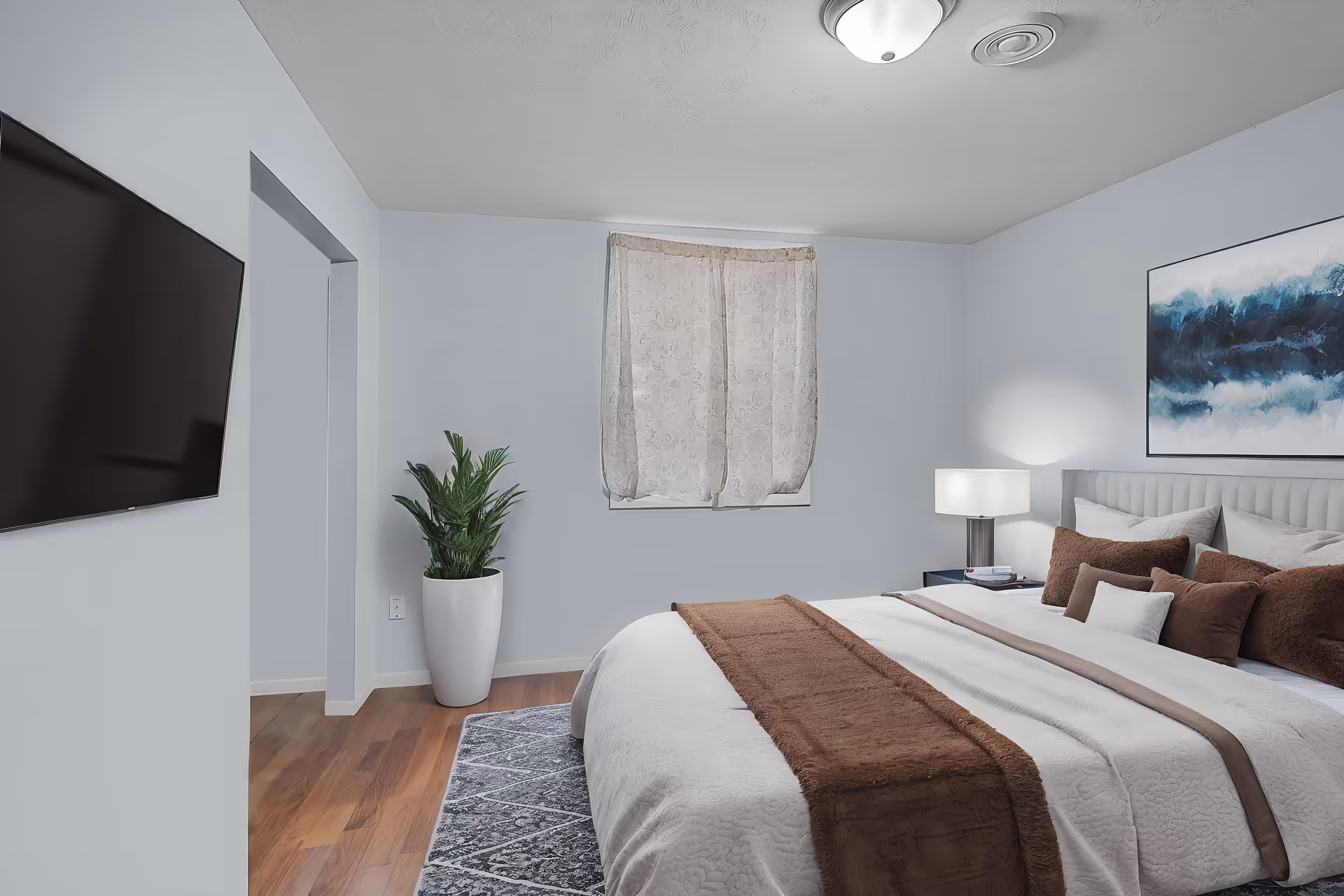 A tidy bedroom featuring a large bed with white and brown linens, a bedside lamp and table, a potted plant, wall-mounted TV, and a curtained window.