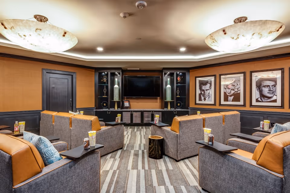 Cozy media/theater room with upholstered recliner-style seats facing a wall-mounted TV, framed portraits on the wall, and warm overhead lighting.