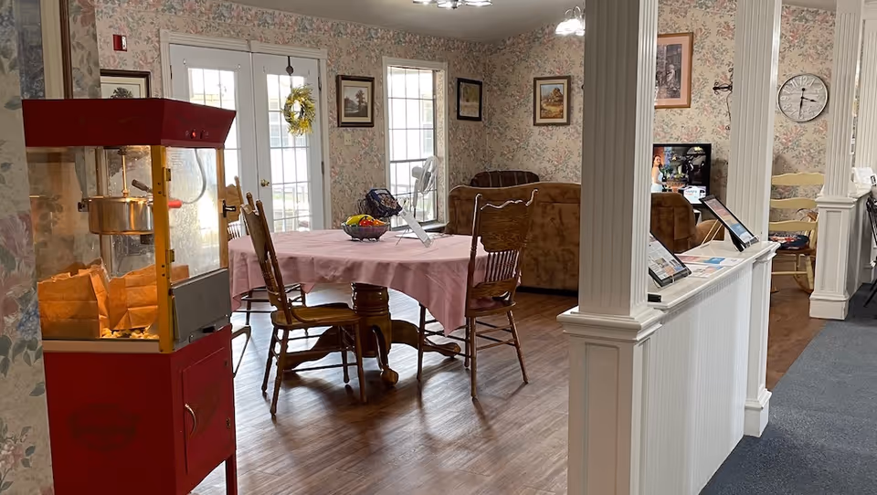 Communal senior living dining area with a pink-clothed table and wooden chairs, a popcorn machine to the left, and a seating area with brochures near white columns.