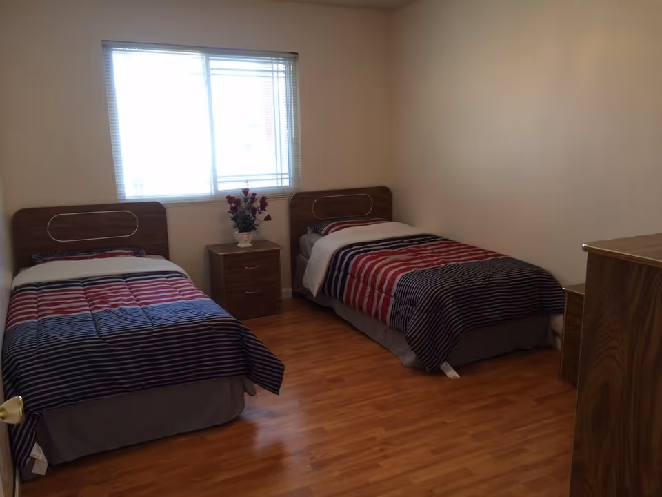 A simple bedroom with two single beds, each with striped bedding in shades of red, black, and white. Between the beds is a small wooden nightstand with a vase of red flowers. A window with blinds is centered on the wall behind the beds, and the floor is wooden. There is also a wooden dresser on the right side of the room.