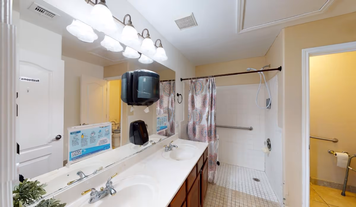 A bathroom with a double sink vanity, large mirror, and overhead lighting. There is a shower area with a colorful patterned shower curtain and grab bars. To the right, there is an open door leading to a restroom with a toilet and grab bars. A paper towel dispenser and soap dispenser are mounted on the wall above the sinks.