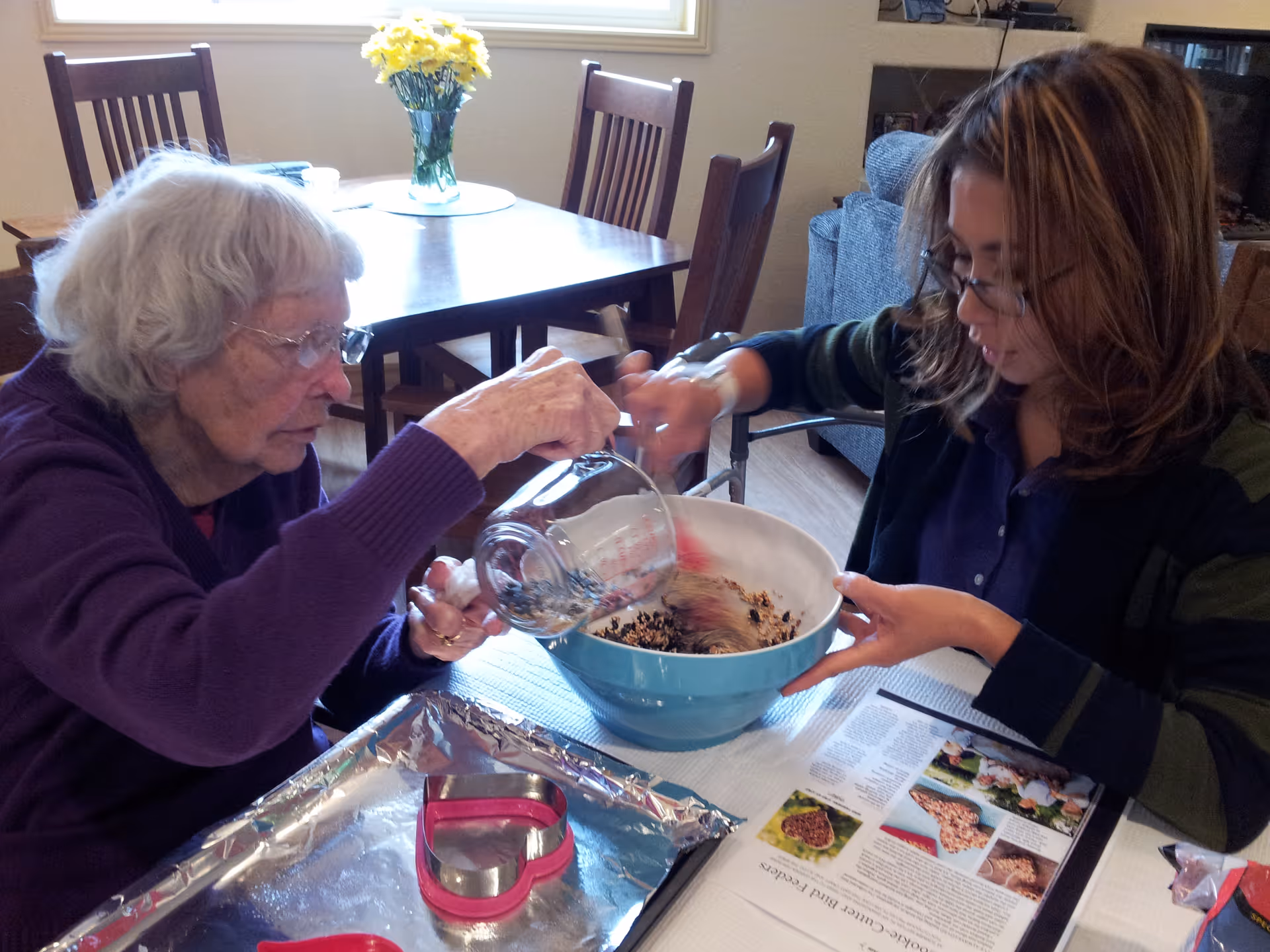Two women mixing ingredients in a large bowl at a dining table with baking tools and a vase of yellow flowers on a nearby table.