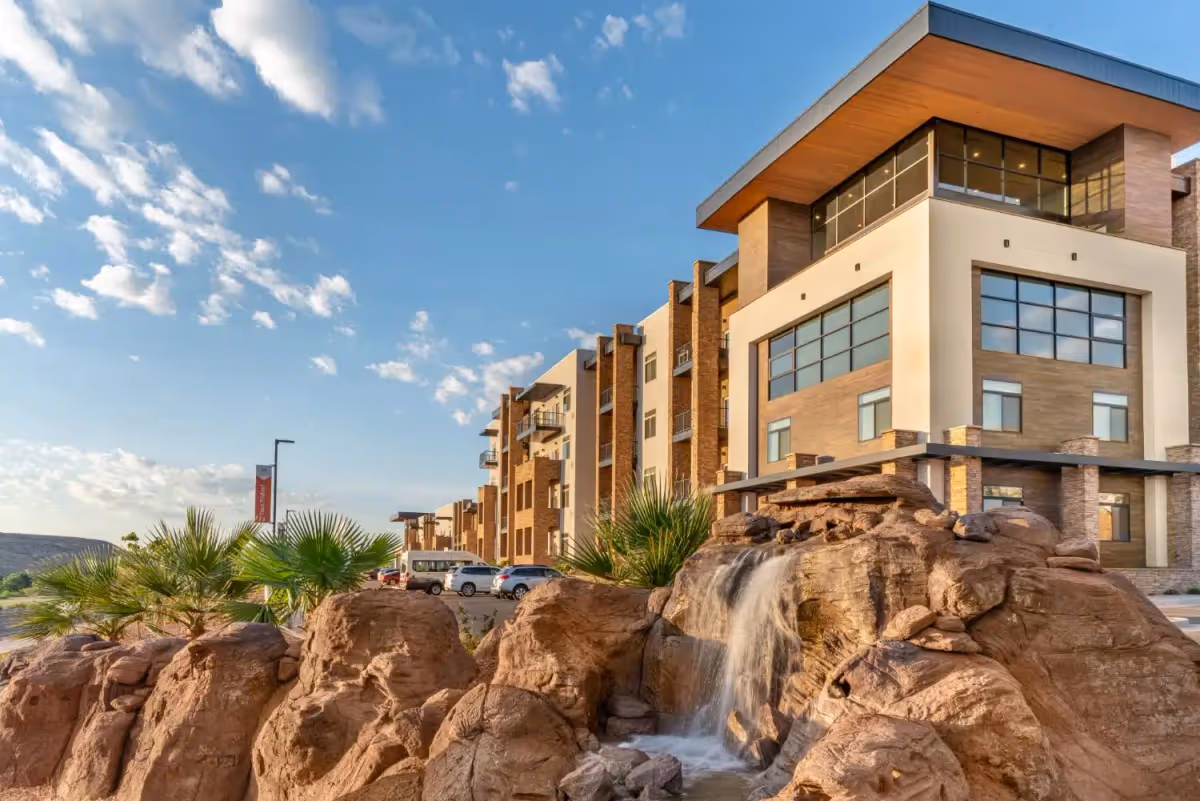 Exterior view of a modern multi-story building with large windows and balconies, surrounded by desert landscaping including rocks, palm plants, and a small waterfall feature under a partly cloudy blue sky.