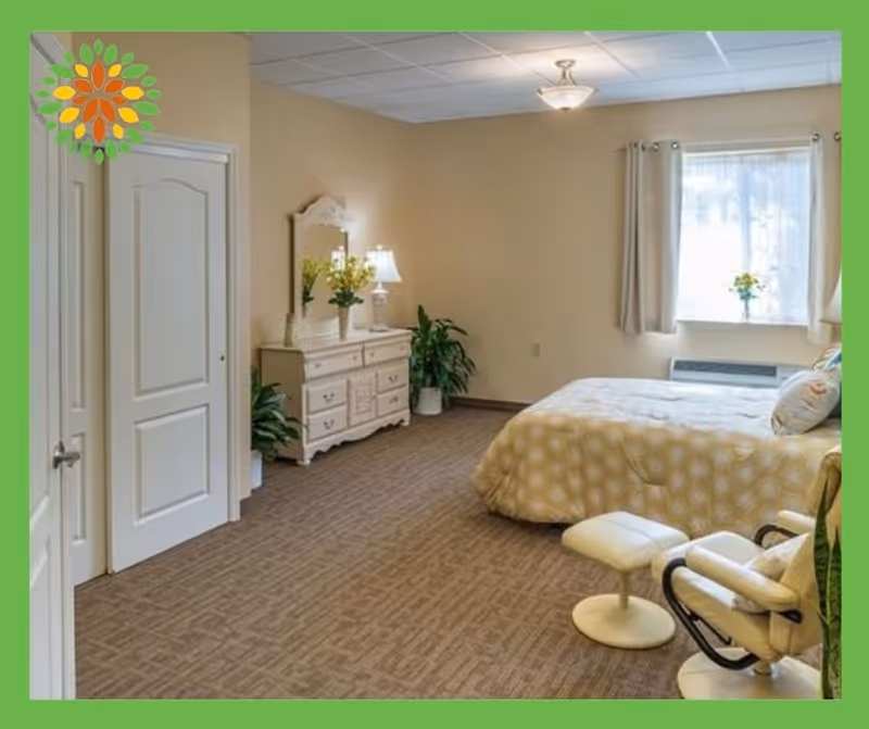 A cozy bedroom with a large bed covered in a yellow patterned comforter, a white dresser with a mirror and a lamp, potted plants, a window with light curtains, and a comfortable chair with a matching footrest.