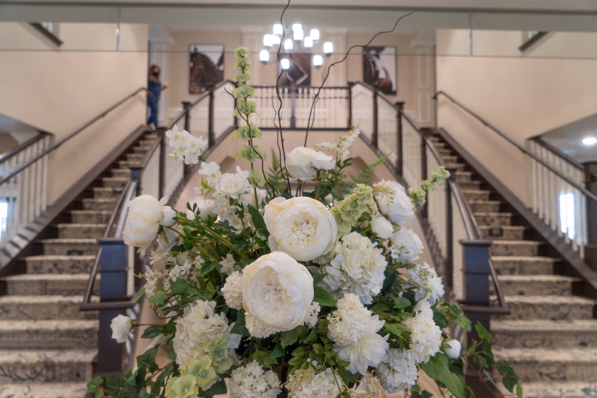 A large floral arrangement with white flowers and green leaves is prominently displayed in the foreground of an elegant interior space featuring a grand staircase with carpeted steps and dark wooden railings. A chandelier hangs from the ceiling above the staircase, and a person is visible standing on the left side of the stairs.