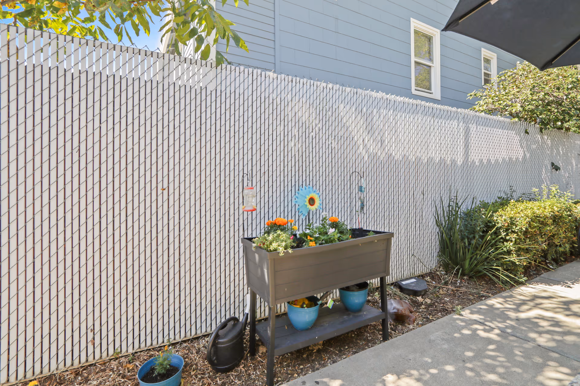 Outdoor garden area with a raised planter box containing flowers and plants, a watering can, and two blue pots underneath. The area is next to a white chain-link fence with a blue house in the background and some greenery along the side.