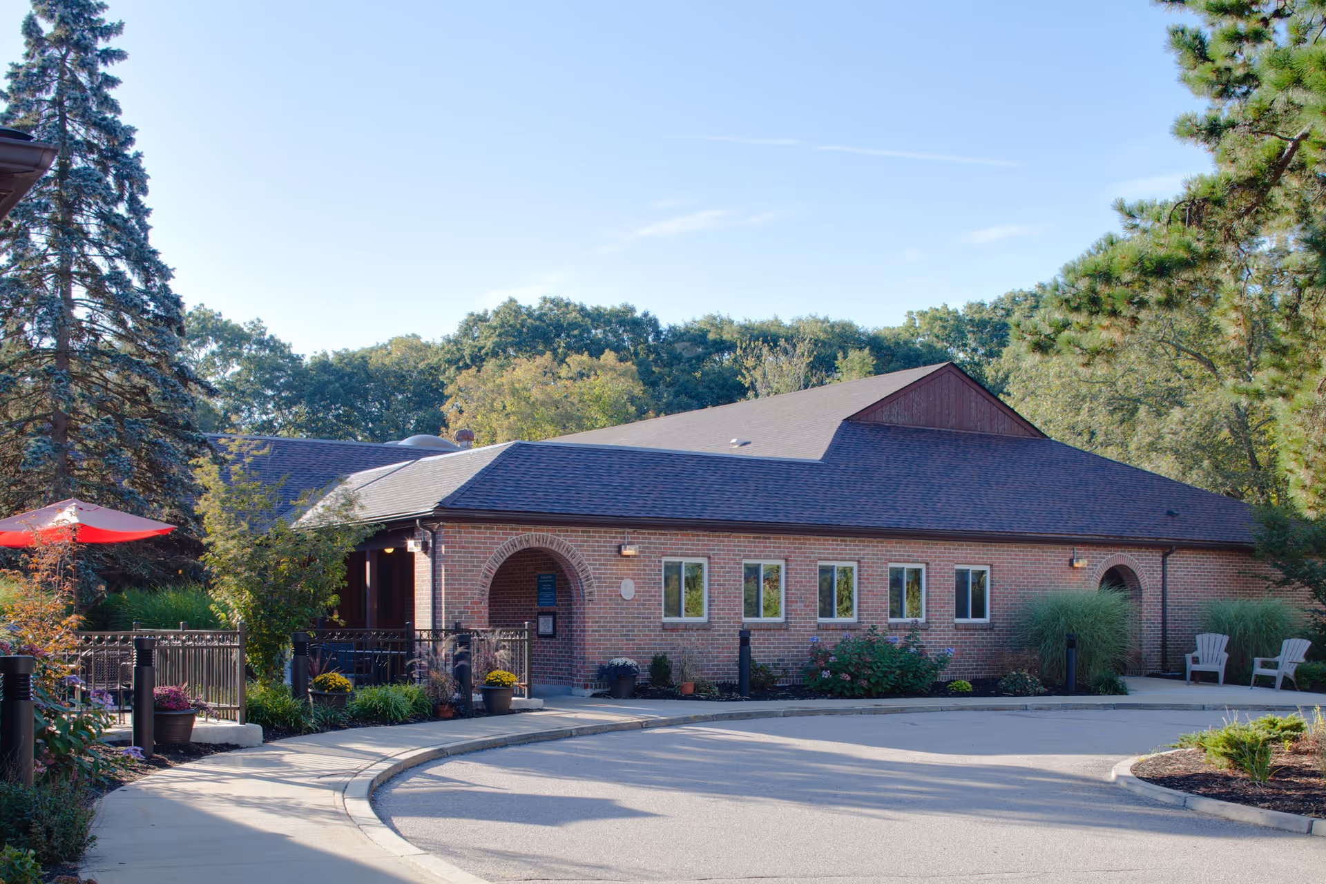 Exterior view of a single-story brick building with a dark shingled roof, surrounded by trees and landscaping. There is a curved driveway in front, a red umbrella on the left side, and two white chairs on the right side near some tall grass.