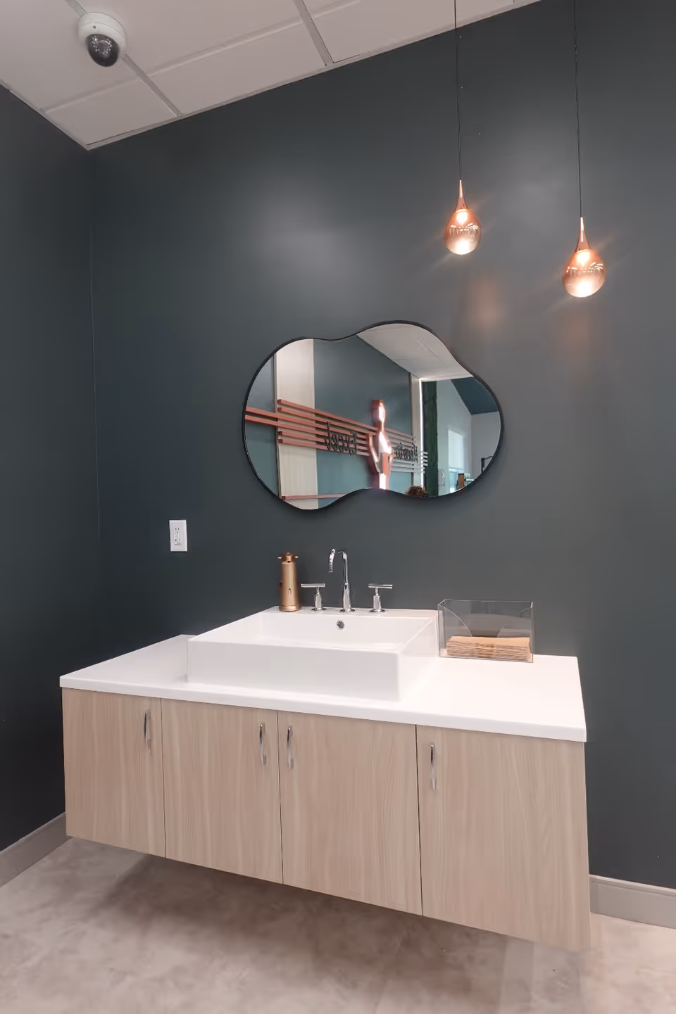 Modern bathroom vanity with a white rectangular vessel sink, an irregular oval mirror, and two pendant lights on a dark green wall.