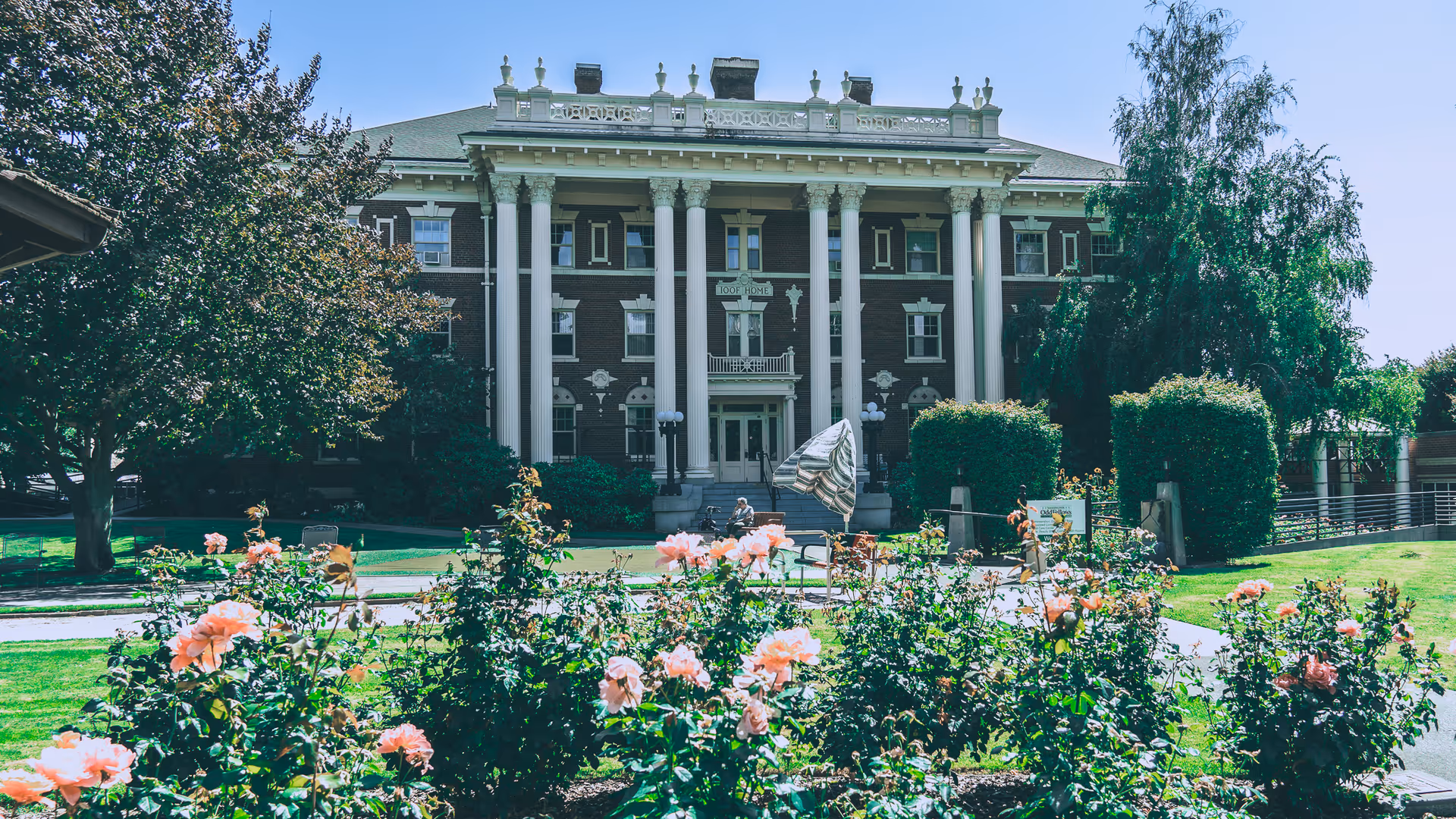 Front exterior view of the Washington Odd Fellows Home, a large historic building with tall white columns and decorative architectural details. In the foreground, there is a garden with blooming pink flowers and green bushes, along with a pathway leading to the entrance.