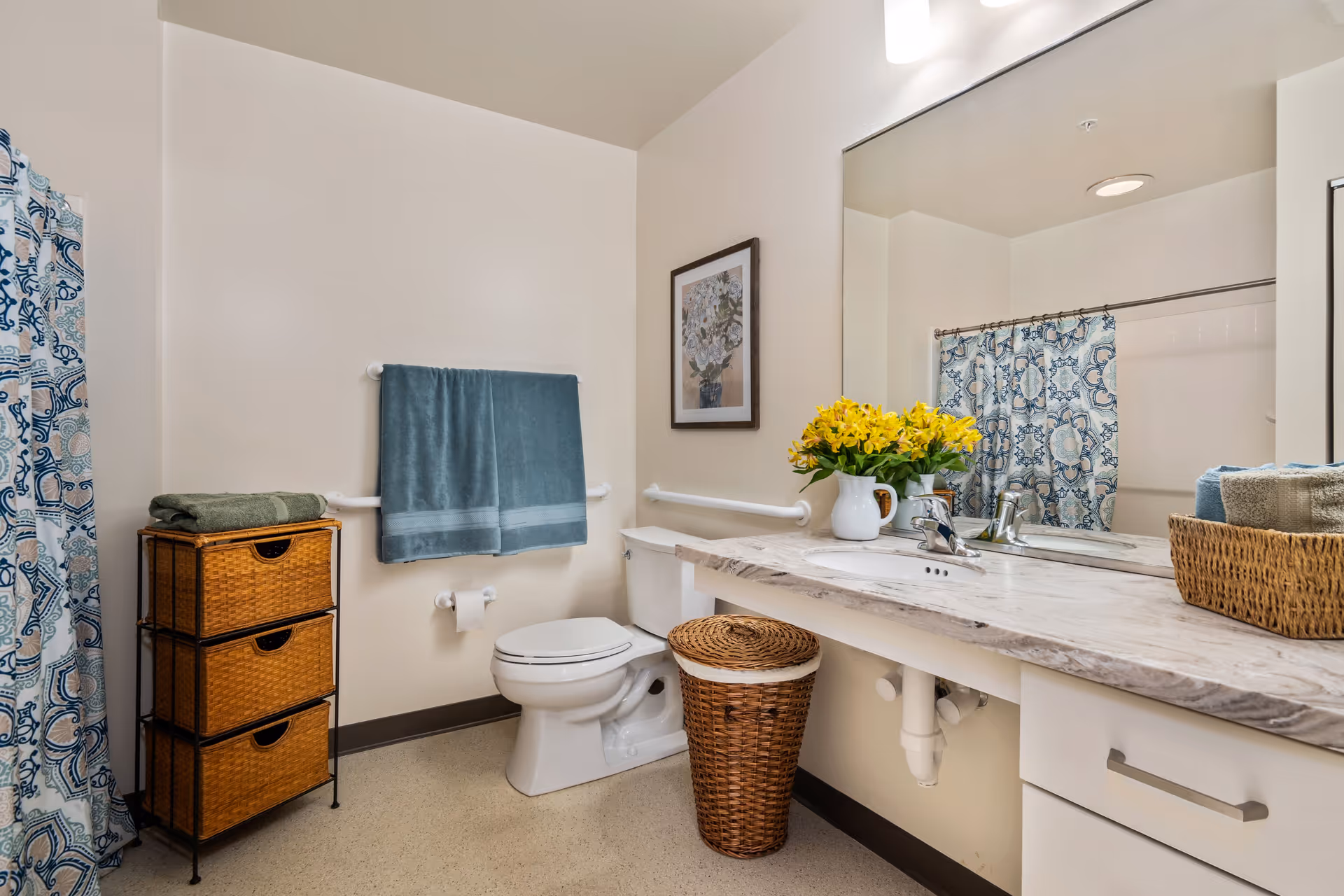A clean and well-lit bathroom featuring a toilet, a large mirror above a marble countertop with a sink, a wicker laundry basket, a small wicker drawer unit, and a towel rack with a blue towel. There is a shower with a blue and white patterned curtain and a framed floral picture on the wall.