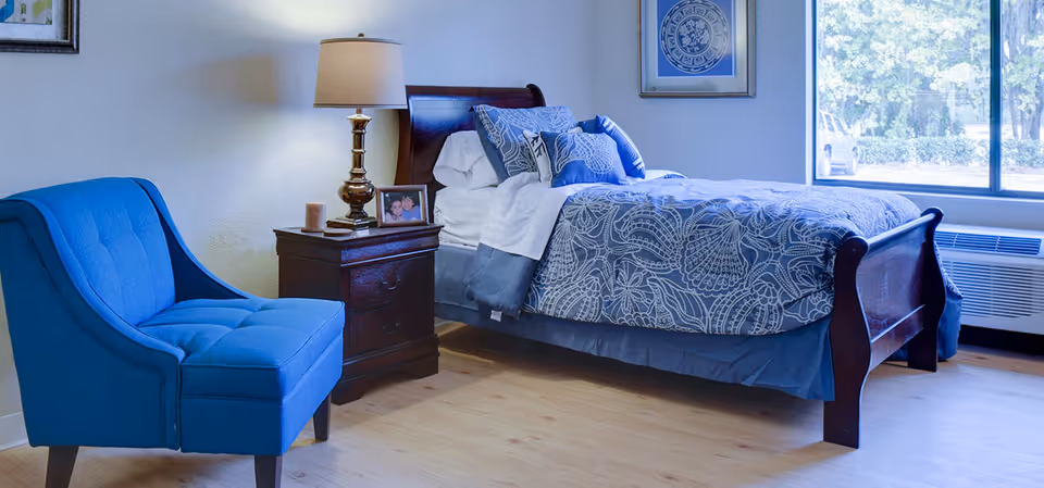 Well-lit bedroom with a wooden bed dressed in blue patterned bedding, a blue upholstered chair, a nightstand with a lamp and framed photo, and a large window.