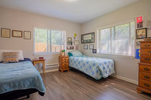 A bright bedroom with two single beds, each with blue bedding. The room has two large windows with blinds allowing natural light to enter. Between the beds is a small wooden nightstand with a lamp and some items on it. The walls are decorated with framed certificates, pictures, and artwork. There is a wooden dresser on the right side of the room. The floor is a wood laminate.