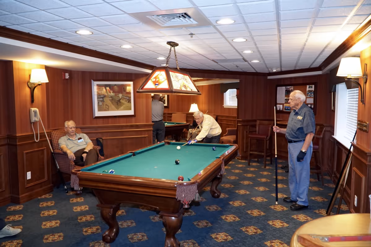 A cozy game room with wood-paneled walls and patterned carpet featuring a pool table in the center. Three elderly men are present; one is sitting in a chair, another is leaning over the pool table preparing to take a shot, and the third is standing holding a pool cue. The room is warmly lit with wall sconces and a stained glass hanging light above the pool table.