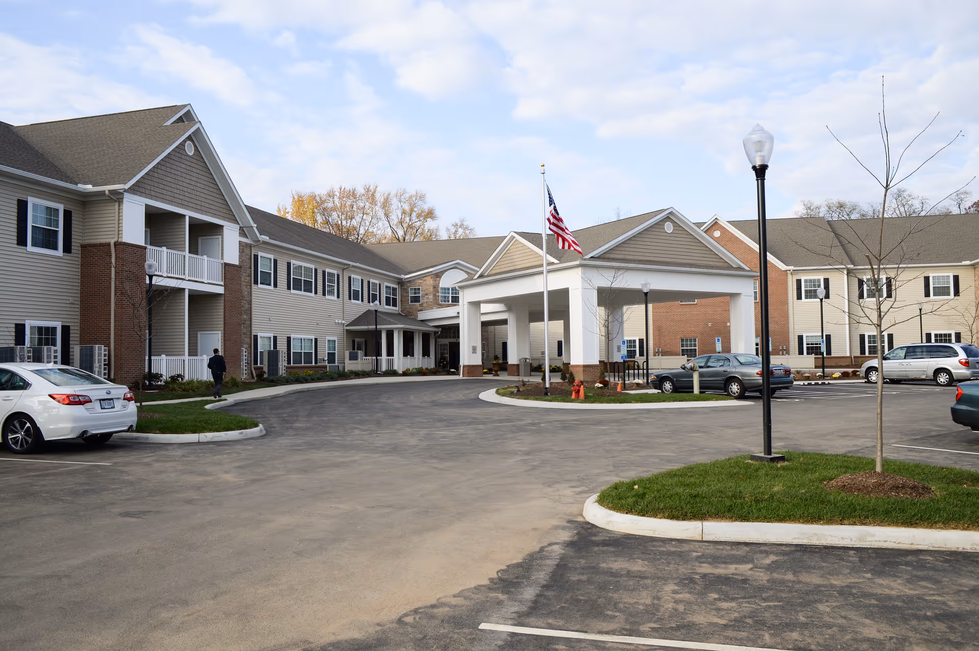 Exterior view of Danbury Tallmadge senior living facility showing a two-story building with beige siding, brick accents, multiple windows, and a covered entrance with an American flag on a flagpole. Several cars are parked in the parking lot, and there are street lamps and small landscaped areas with grass and young trees.