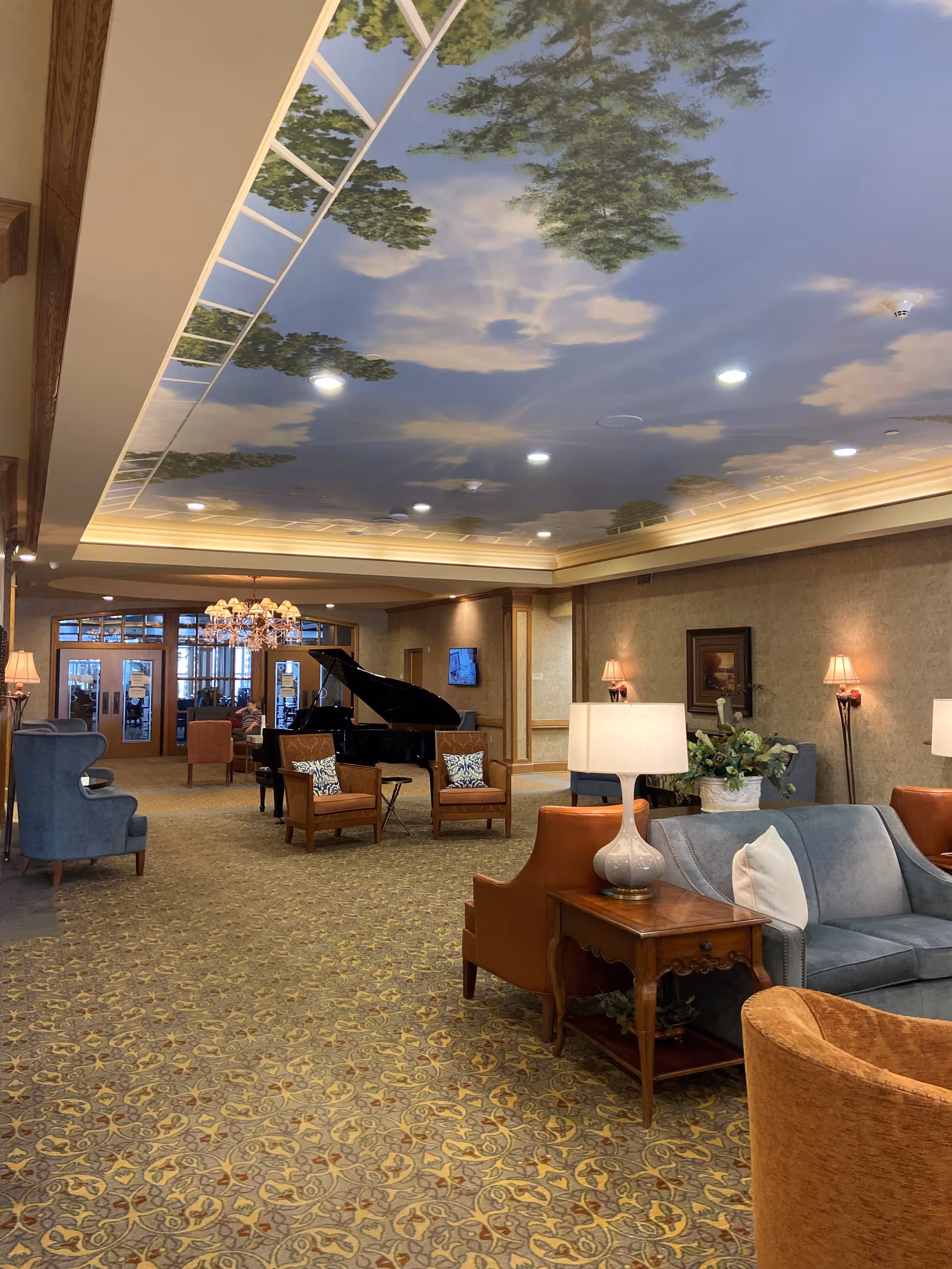 Elegant communal lounge with sofas and chairs centered around a grand piano under a painted sky ceiling.