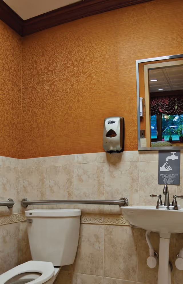 A clean and accessible bathroom with beige tiled walls and an orange patterned wallpaper above. The bathroom features a white toilet with grab bars on both sides, a white pedestal sink with a silver faucet, a wall-mounted Gojo soap dispenser, and a mirror reflecting part of a window with curtains. There is a sign below the mirror reminding employees to wash hands before returning to work.