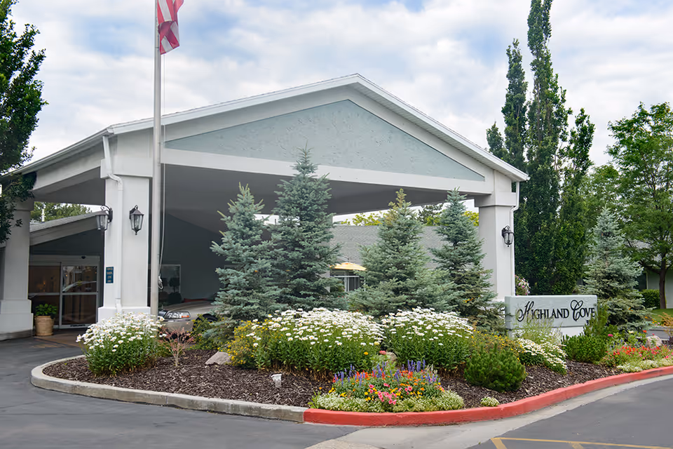 Entrance of Highland Cove Retirement facility with a covered driveway, landscaped garden featuring evergreen trees and colorful flowers, an American flag on a flagpole, and a sign displaying the facility's name.